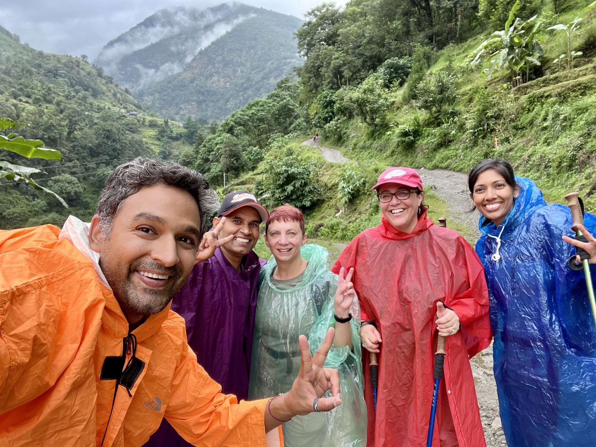Two men and three women wearing rain ponchos pose for a selfie with a dirt trail and green mountains behind them.