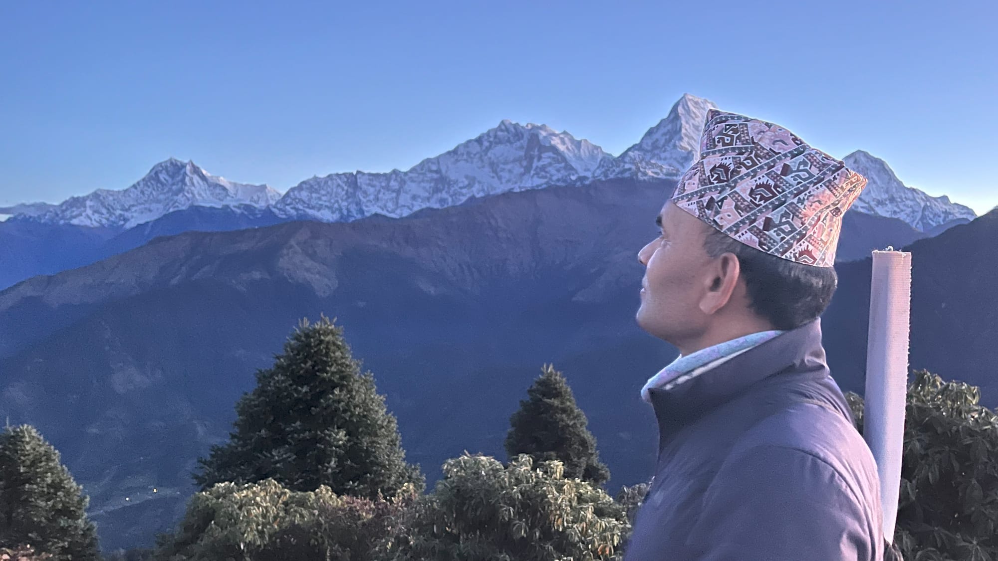 Nepalese man wearing traditional Dhaka Topi hat looks out over tree tops at snow-capped mountains in the Annapurna range of the Himalayas.